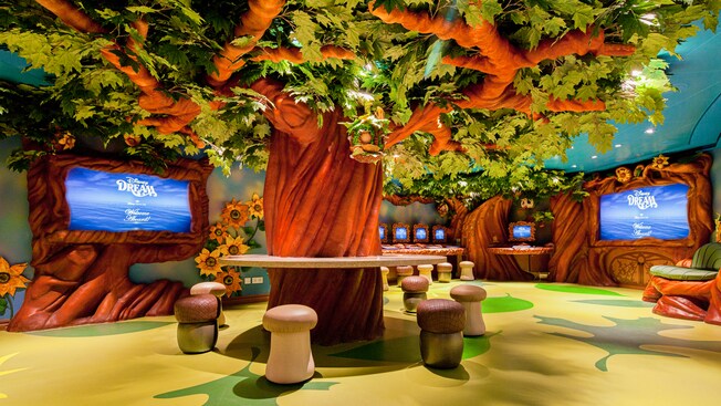 A room with a giant tree is surrounded by acorn and mushroom shaped stools, video screens and computers