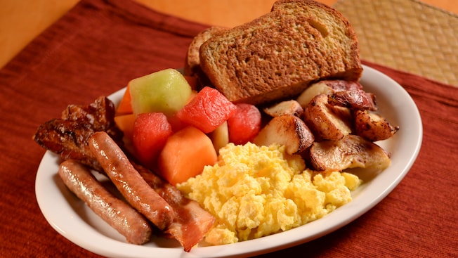 A plate topped with bacon, sausage links, scrambled eggs, potato wedges, toast and chunks of fruit