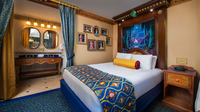 Royal bed with tall regal headboard, framed Disney princesses on wall, curtained bathroom with vanity