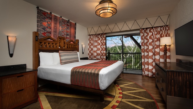 Interior of a room with wood flooring, a dresser, 2 nightstands, a wall mounted flat screen TV, a king bed, curtains and a sliding glass door with balcony access