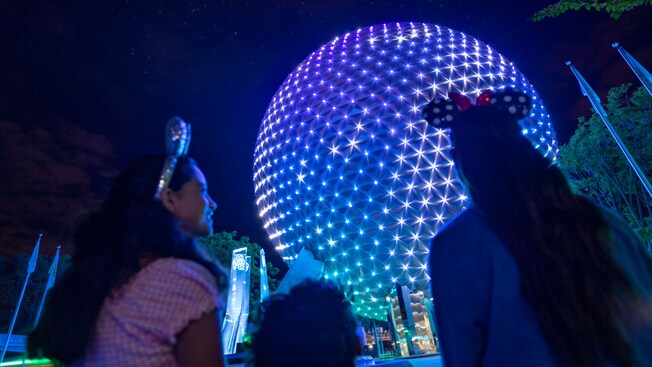 Three girls, two of them in Minnie Mouse ears, look up at the decorative lights on Spaceship Earth