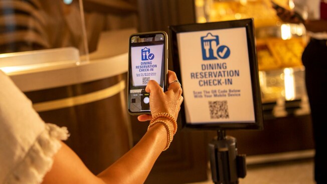 A Walt Disney World Guest using her phone camera to scan a sign featuring a QR code and text that reads ‘Dining Reservation Check In. Scan the QR code below with your mobile device’