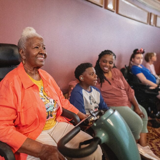 A woman in a scooter smiles while watching a show, with a boy and another woman sitting beside her.