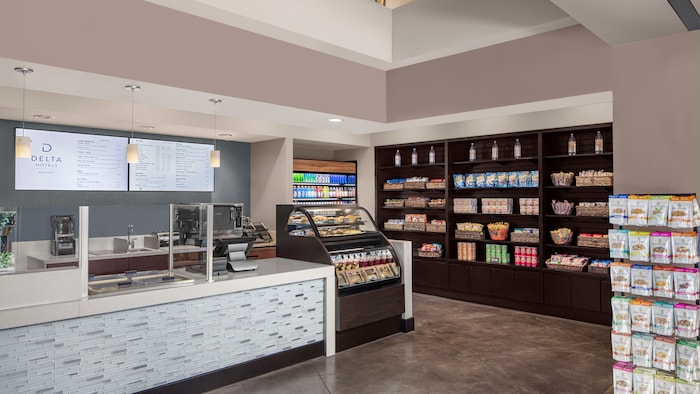 The hotel convenience store features a service counter, refrigerated cases, freestanding racks and shelving units filled with boxed meals, snacks and beverages