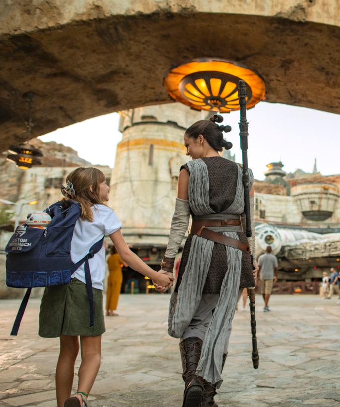 A girl with a backpack holds Rey's hand as they walk through Star Wars Galaxy's Edge