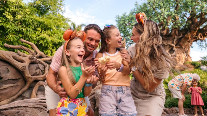 A family of 4 shares a joyful moment with a Mickey-shaped treat in front of the Tree of Life at Disney’s Animal Kingdom Theme Park