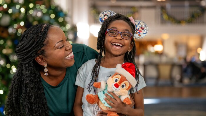 A young girl wearing Minnie Mouse ears, holding a Dale Christmas plush, laughing with her mother, at a Disney World Resort hotel with seasonal decorations
