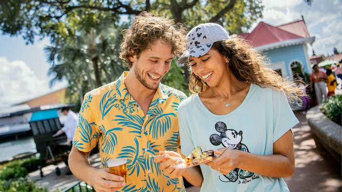 Una pareja camina junto a la laguna de World Showcase mientras disfruta de un snack y una cerveza en EPCOT International Food & Wine Festival