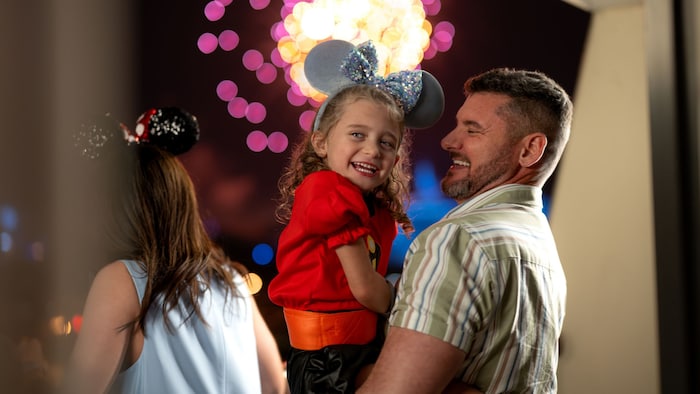 A father holds his daughter in his arms while they smile as fireworks burst behind them