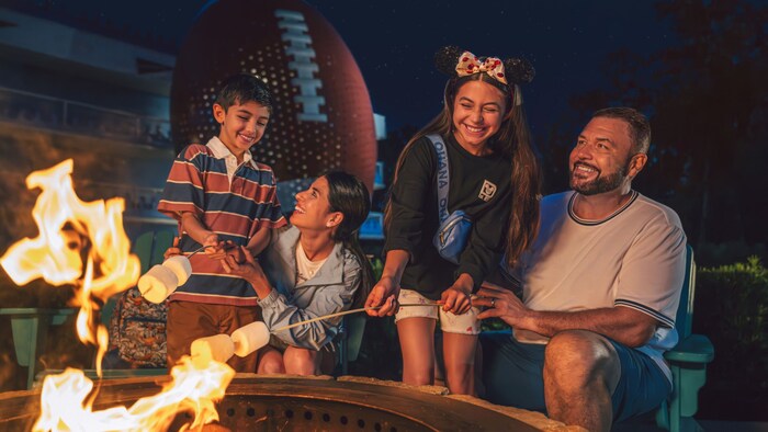 A family of 4 smiles as they roast marshmallows over an open fire at night