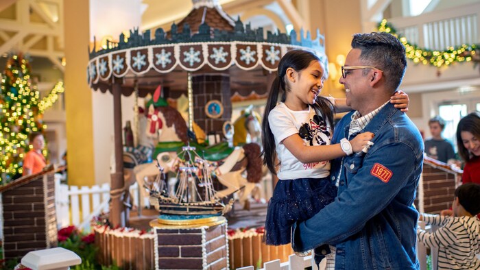 A father holding his young daughter in his arms while standing in front of a holiday carousel at Disney’s Beach Club Resort