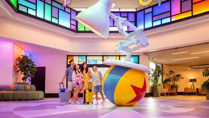 Family with luggage walks past the giant Pixar Ball and Luxo Jr. lamp in the Pixar Place Hotel lobby