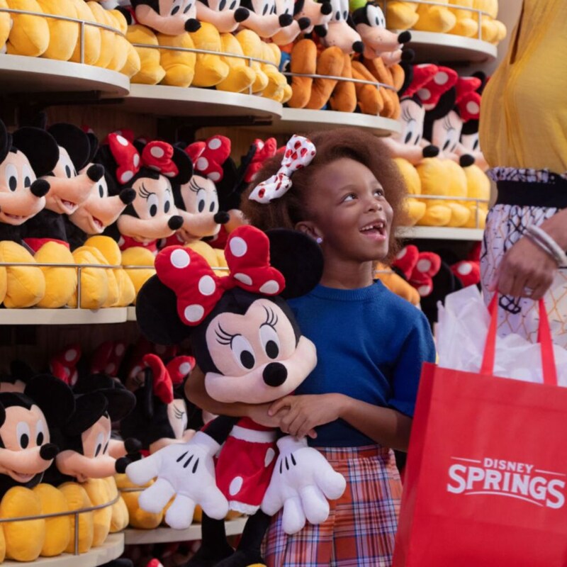 A young girl holds a Minnie Mouse plush in front of a wall of other plushes at Disney Springs