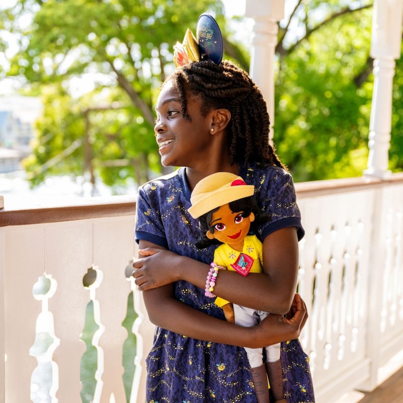 A girl hugging a Tiana plush doll while wearing a Tiana’s Bayou Adventure Ear Headband and a dress featuring Ray from ′The Princess and the Frog′