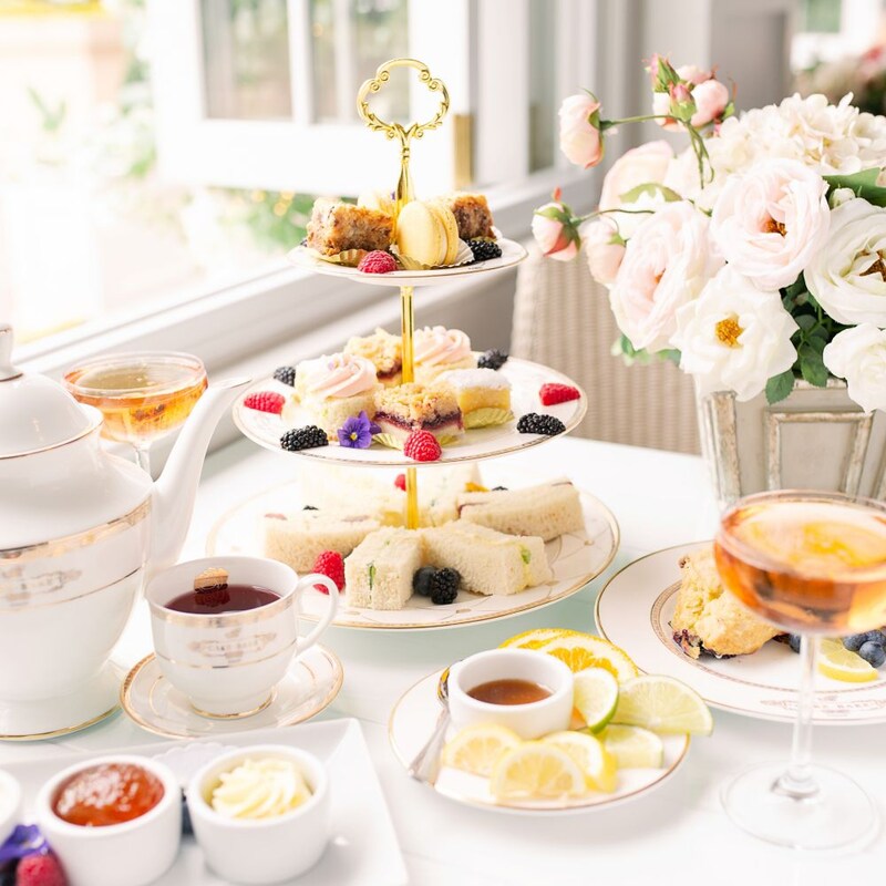 An elegant set up for afternoon tea featuring a tea pot, finger sandwiches, mini desserts and a cocktail