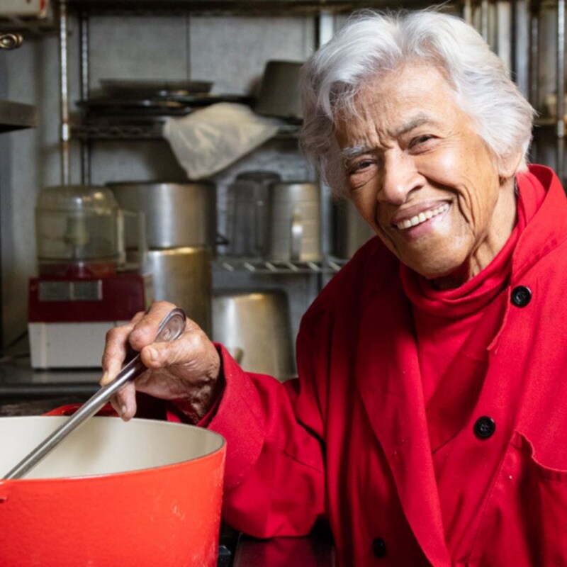 A chef Leah Chase mexendo uma panela grande com uma colher de metal em um fogão