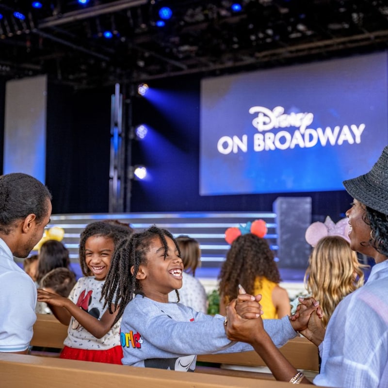A seated family and a projector on stage with the words, ‘Disney on Broadway’