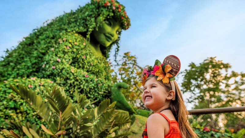 Una niña sonriendo con vestimenta temática de Moana frente a un arbusto artístico de Te Fiti en Journey of Water, inspirado en Moana, en Epcot