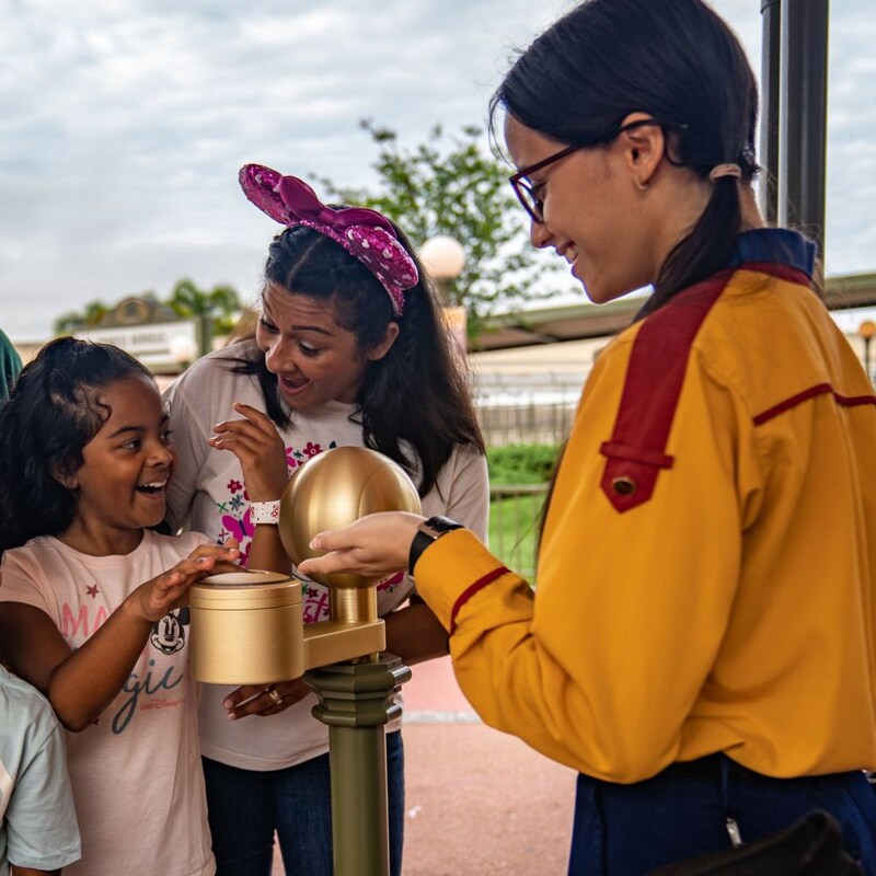 A young Guest uses a Disney band to interact with a park experience while her mom and a Cast Member look on