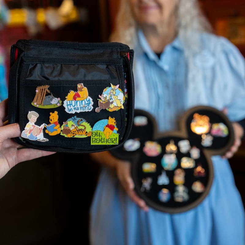 A hand holding a pin trading book with a Cast Member holding a Mickey Mouse shaped pin tray nearby