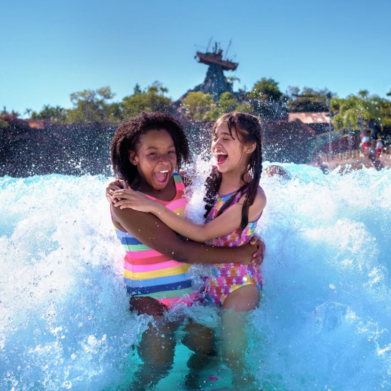 2&nbsp;filles dans une piscine à vagues se tiennent les unes les autres alors qu’une vague déferle derrière elles