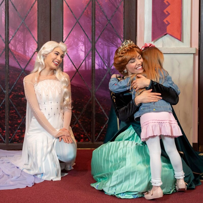 Anna crouches to hug a child as Elsa smiles at them