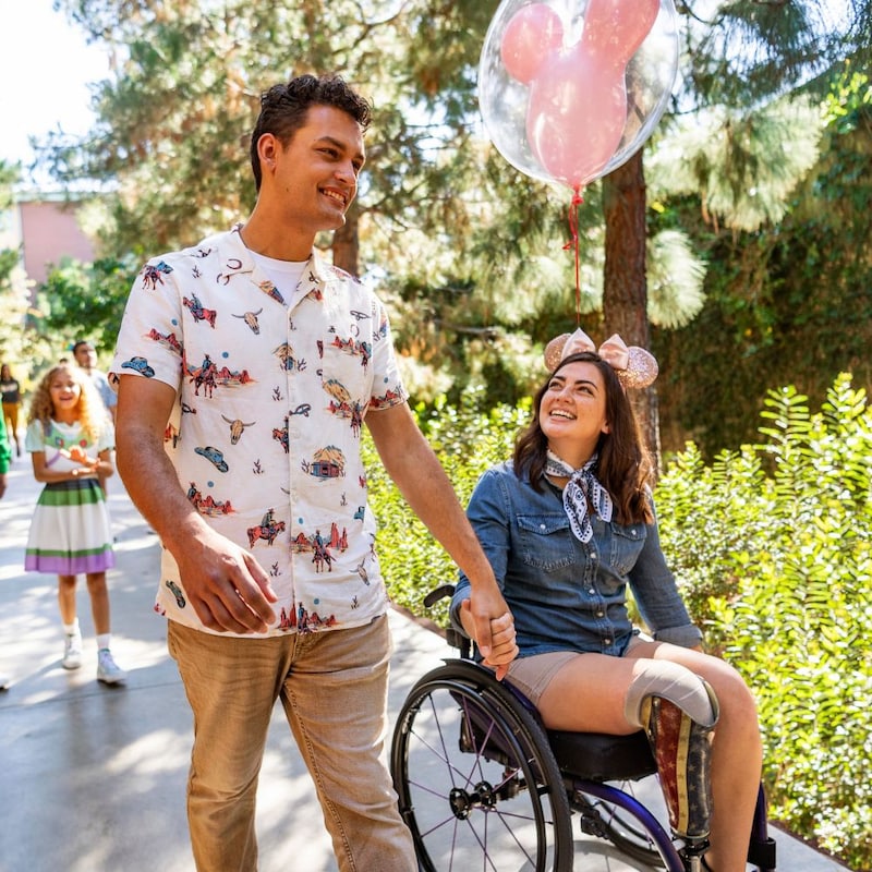 A woman in a wheelchair holds hands with a man while walking in a park