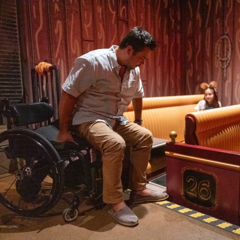 Un hombre se desliza de su silla de ruedas al asiento del vehículo para Mickey and Minnie's Runaway Railway, mientras una niña más joven observa desde el vehículo.