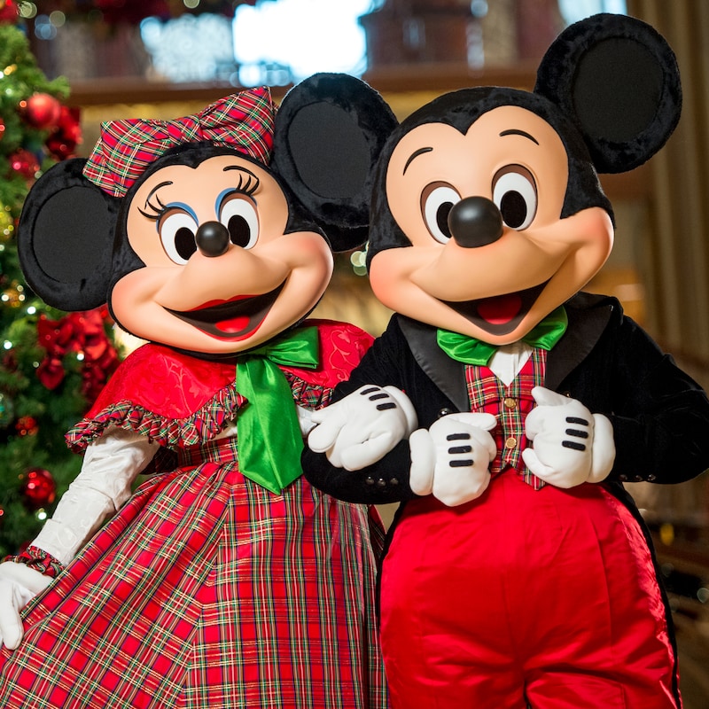 Dressed in holiday attire, Mickey Mouse and Minnie Mouse pose together in front of a Christmas tree