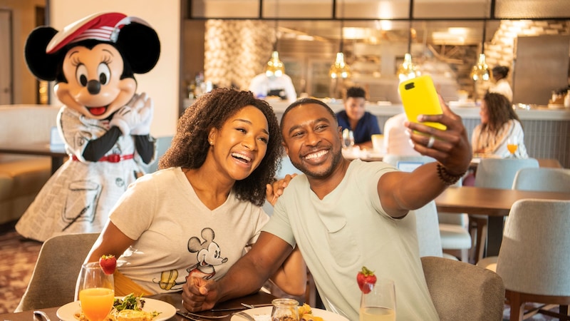 A man and woman sitting at a table for breakfast at Topolino’s Terrace Flavors of the Riviera, taking a selfie together with Minnie Mouse in the background