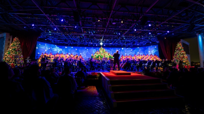 A conductor leading a large choir and orchestra on a festive holiday stage flanked by decorated Christmas trees