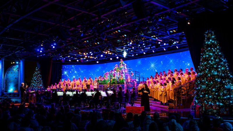 A conductor leading a large choir and orchestra on a festive holiday stage flanked by decorated Christmas trees