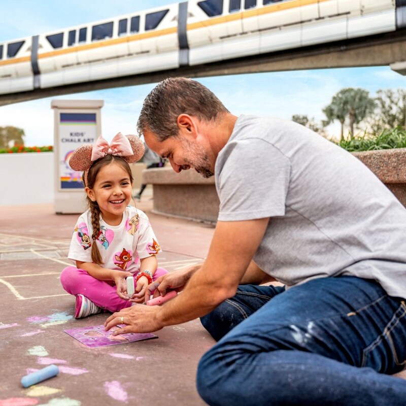 Un padre y sus 2 hijas dibujando con tiza en el suelo cerca del monorriel de Walt Disney World