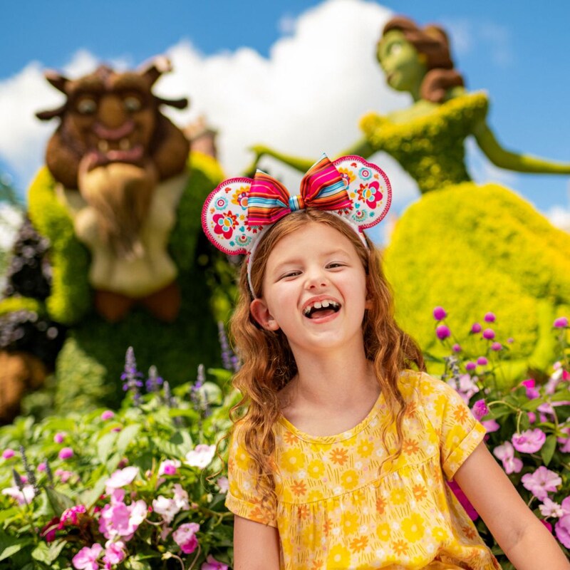 Una niña con una diadema de orejas de Minnie Mouse sonríe cerca de un arbusto artístico de Beauty and the Beast en un jardín botánico