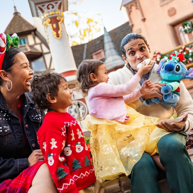Un papá sostiene un peluche de Stitch y su hija le ofrece un bocado de su sándwich mientras otros miembros de la familia ven