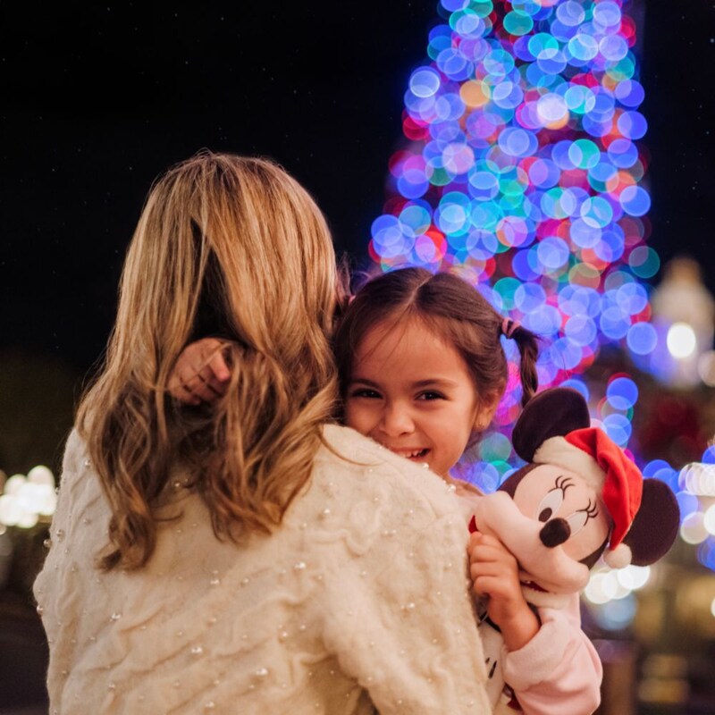 Una niña sonriente sostiene un peluche de Minnie Mouse mientras su mamá la carga