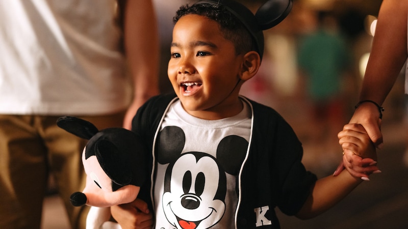  A young boy in a Mickey Mouse ear hat holding a Mickey Mouse plush in one hand and an adult's hand in the other