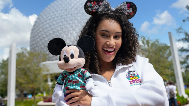Guest in a Mickey Mouse pullover and ear headband carrying a Mickey Mouse plush from the Mickey and Friends 2026 collection at Spaceship Earth in Epcot