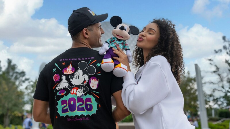 A man showing off his Mickey and Friends 2026 collection shirt and a woman holding a Mickey Mouse plush at Spaceship Earth in Epcot
