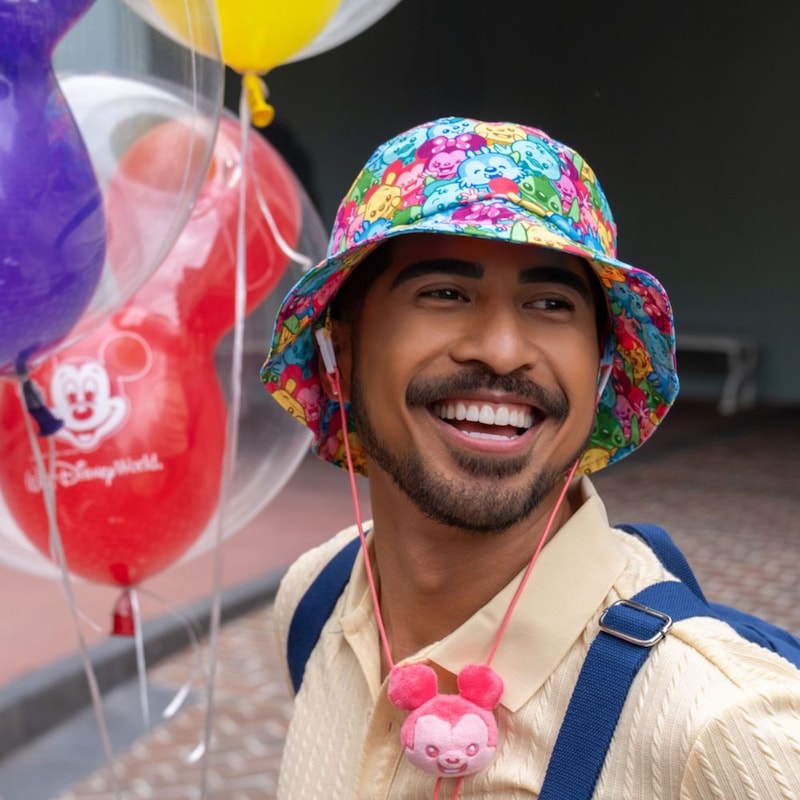A man wearing a bucket hat featuring Disney characters while holding Mickey Mouse balloons