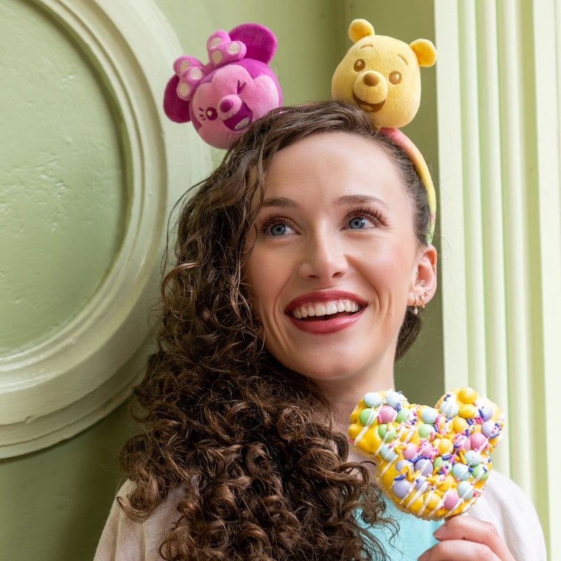 A woman wearing a Create Your Own Headband featuring Minnie Mouse and Winnie the Pooh while holding a Mickey shaped treat