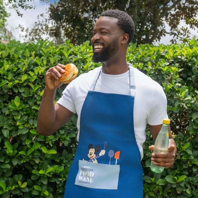 A Guest wearing a blue Mickey Mouse Food and Wine Festival apron while holding a snack and bottle