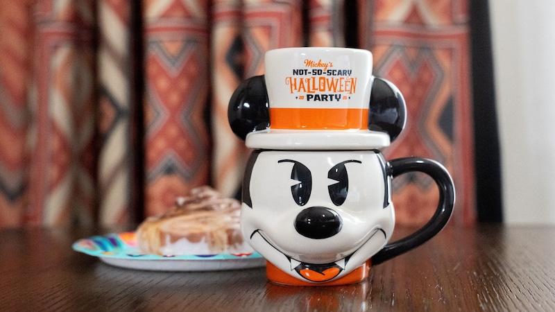 A Mickey Mouse shaped ceramic mug reading Mickey's Not So Scary Halloween Party on a wooden table beside a pastry