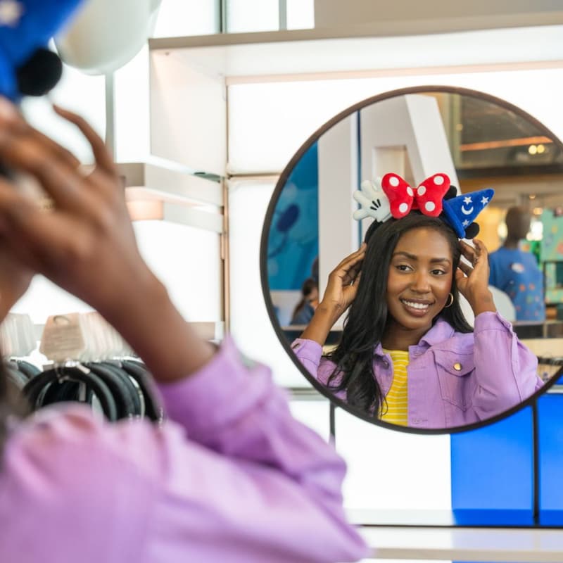 A Guest looking at her reflection in a mirror while trying on a Create Your Own Headband featuring Fantasia and Minnie Mouse plush elements