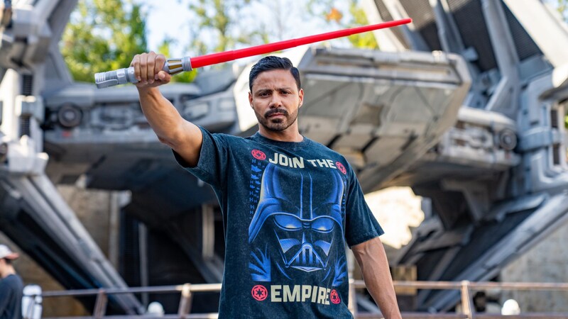 A man holding up a lightsaber toy and wearing a Darth Vader T shirt at Star Wars Galaxy’s Edge