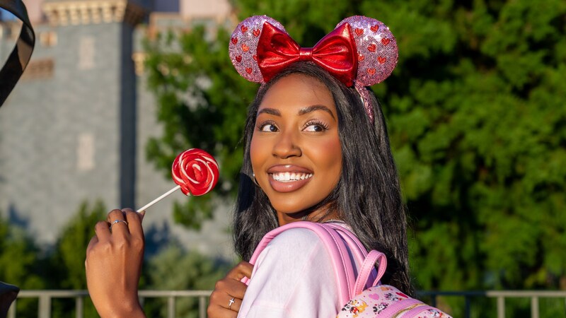 A Guest holding a lollipop and wearing a heart covered Minnie Mouse ear headband