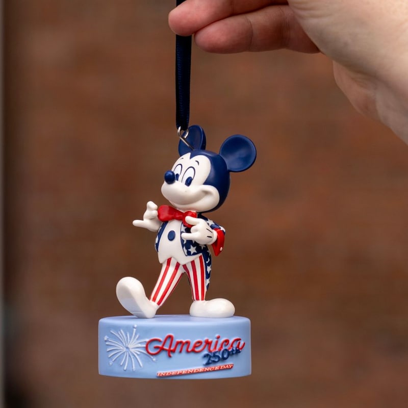 A hand holding a small Mickey Mouse ornament that says ‘America 250th'