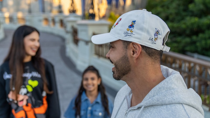 Two Guests standing near a man wearing a baseball hat featuring different Disney characters