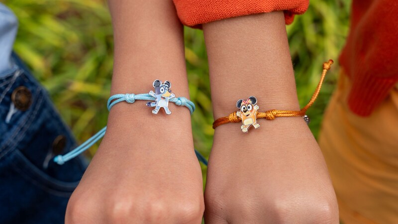 Two wrists wearing string bracelets featuring charms of Bluey and Bingo