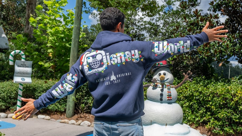 A man wearing a Walt Disney World holiday hoodie stretching his arms out while standing in a mini golf course